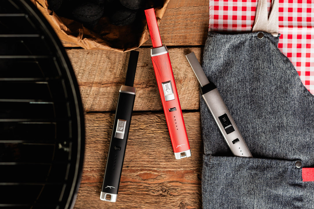 Three Sizzle Pitmaster USB rechargeable lighters in Black, silver and Redwood color lying beside a barbeque and cooking apron