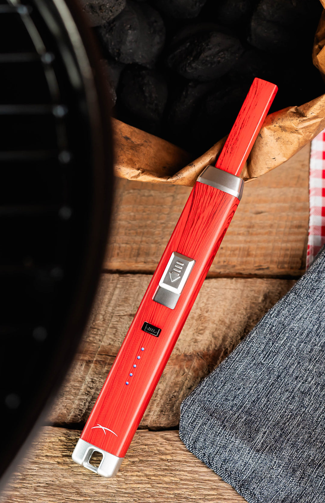 Sizzle Pitmaster USB rechargeable lighter in redwood color beside a bag of charcoals and a barbeque grill
