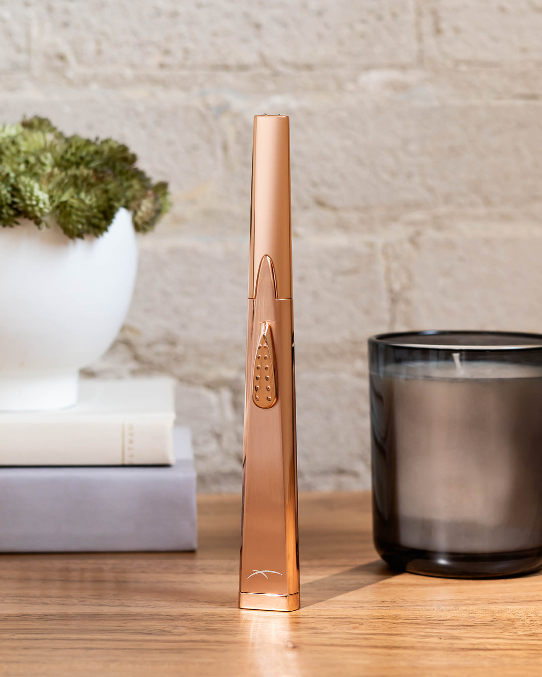 Sizzle Sleek USB rechargeable lighter in rose gold metal casing standing upright on a wooden table with a candle in a glass, a plant in a ceramic jar on two books and a white brick wall staged behind