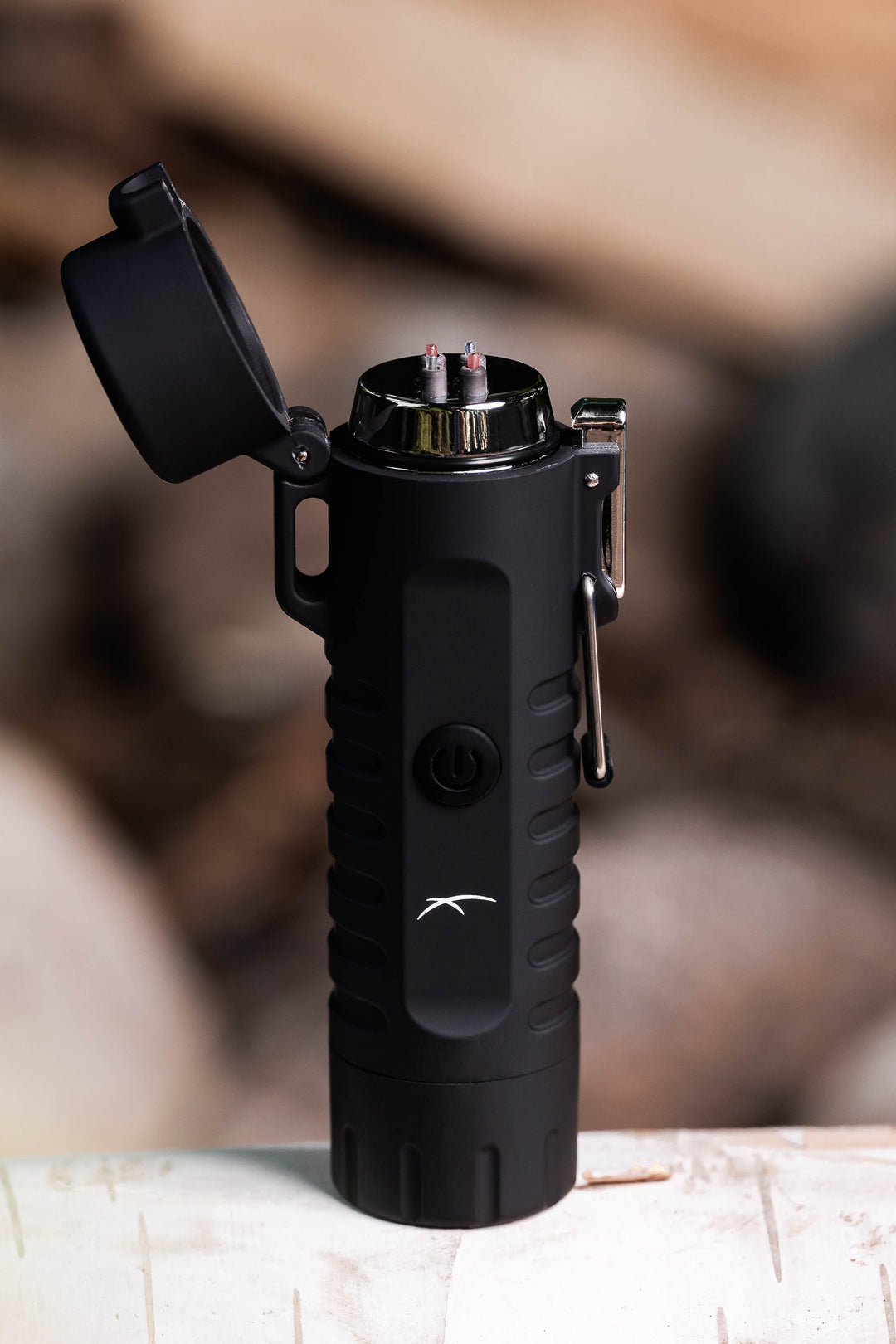 A Sizzle Survival USB rechargeable lighter/flashlight in black waterproof casing standing upright on a birch log with lid open