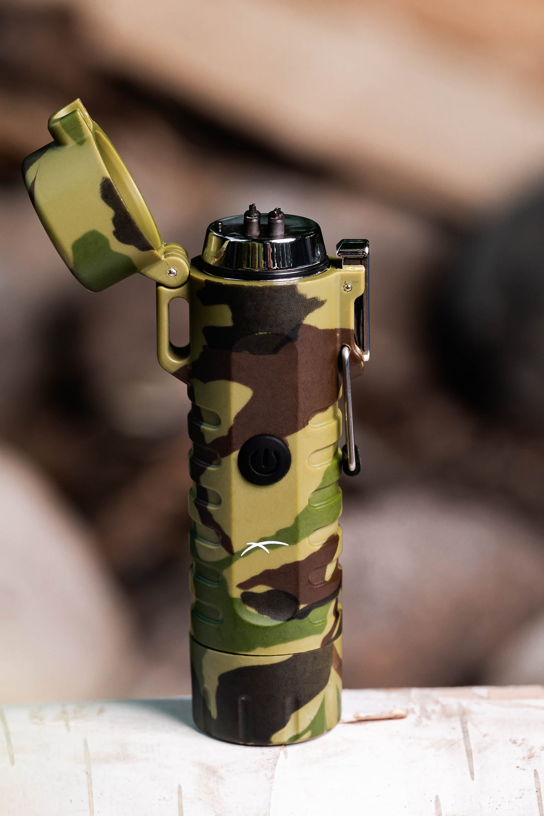A Sizzle Survival USB rechargeable lighter/flashlight in camouflage waterproof casing standing upright on a birch log with lid open 