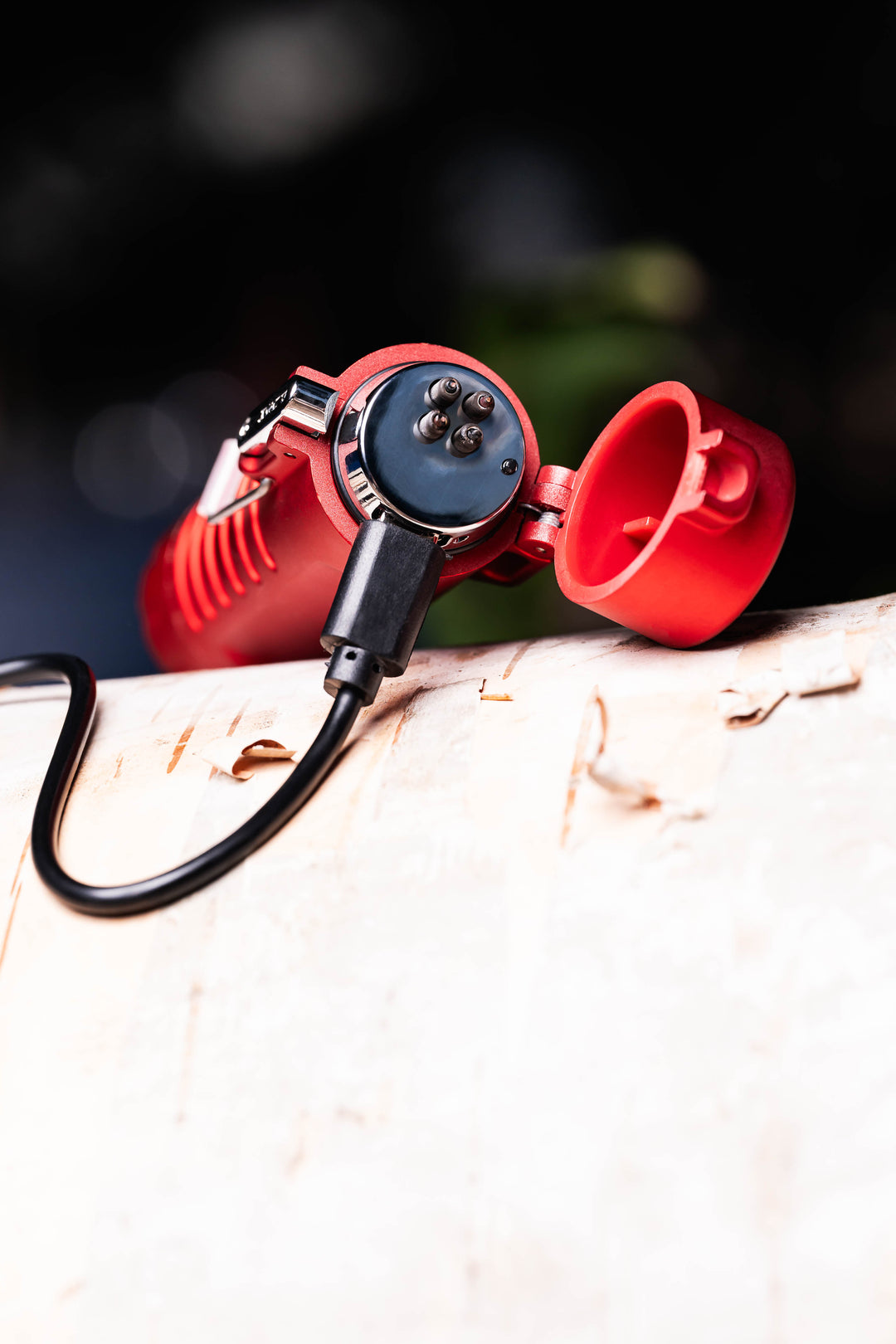 A Sizzle Survival USB rechargeable lighter/flashlight in red casing lying on top of a birch log with lid open and USB charge cable inserted